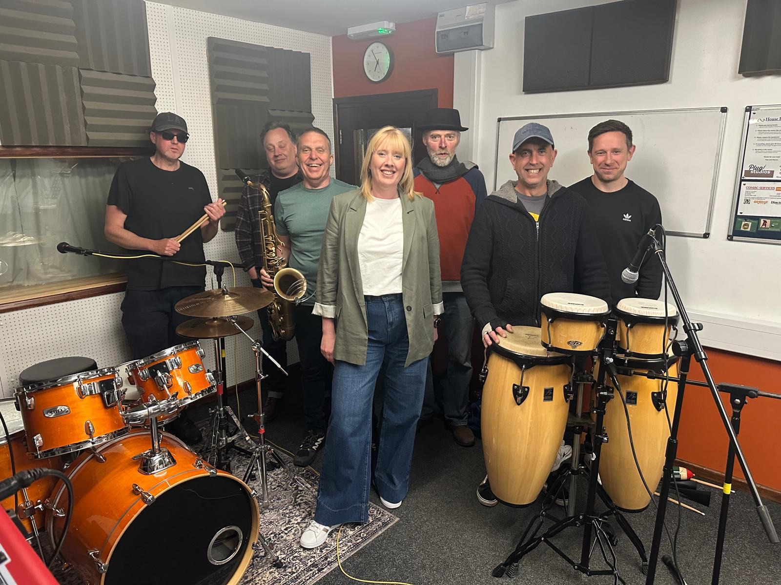 The full band in the rehearsal studio with drums, congas and saxophone